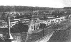 Зернистая, черно-белая фотография показывает несколько одиночных зданий сюжетных рядом с грунтовым дорогой.  Несколько домов и других зданий тянутся в сторону гор на расстоянии.  Труба видна на переднем плане.
